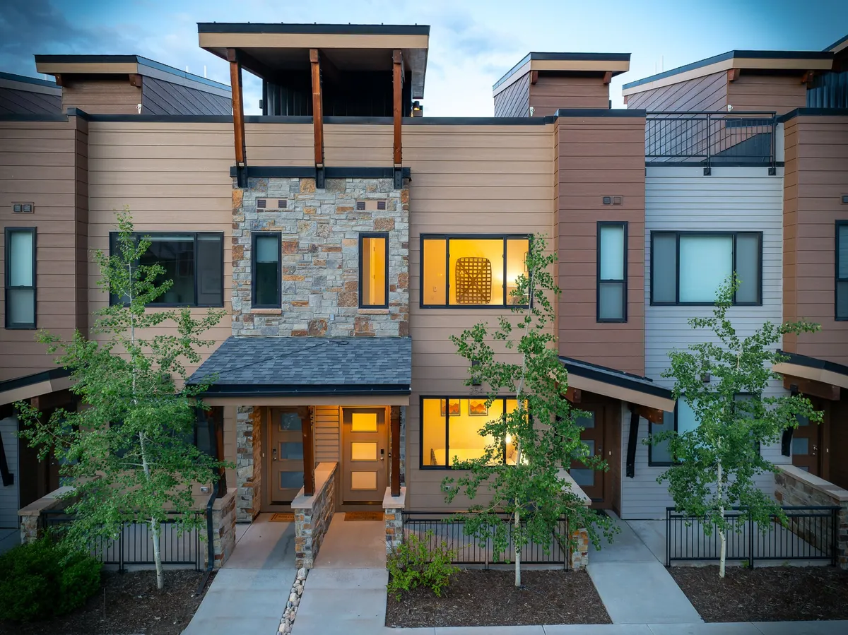 An inviting exterior view of the front entrance with a stone facade and large windows. Warm light glows from inside, suggesting evening activity, while the spacious balcony hints at the opportunity for gatherings and sunset views.