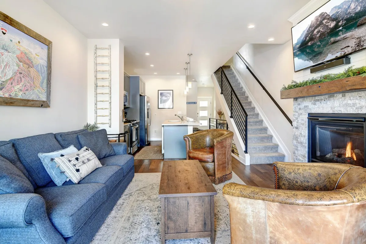 This image provides a wide view of the open-concept great room, showcasing the plush sofa, a glowing fireplace, the adjacent dining area, a staircase, and tasteful artwork. The space feels welcoming and exudes both comfort and style, highlighting areas for relaxing, entertaining, and sharing meals.