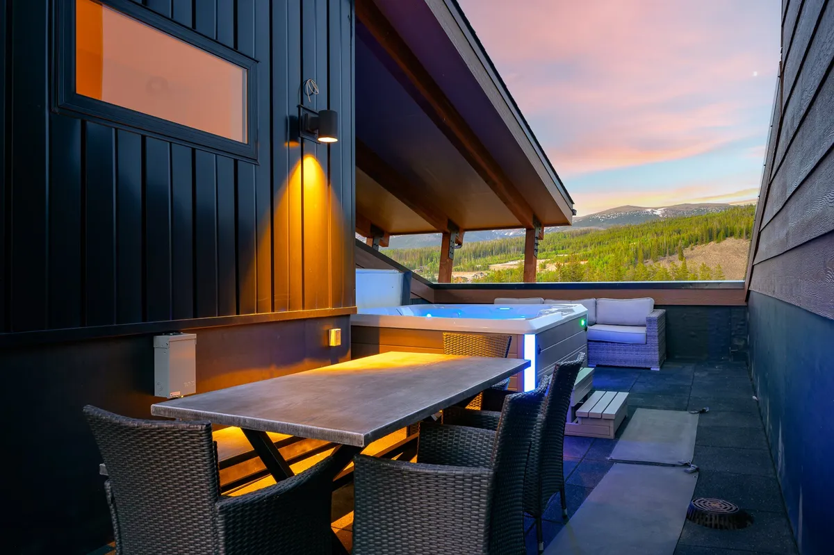 An outdoor space featuring a dining table, bubbling hot tub, and comfortable lounge seating, all under an open sky with sunset hues. The layout invites group gatherings, al fresco meals, and relaxation, with glimpses of the surrounding landscape.