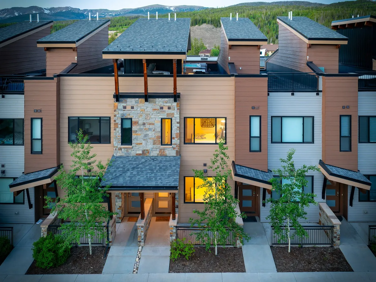 The image showcases a stunning modern villa exterior with a beautiful stone facade, expansive windows that promise mountain views, and aspen trees gracefully lining the entrance walkway. The setting exudes an inviting, upscale mountain retreat vibe, perfect for family gatherings and outdoor relaxation.