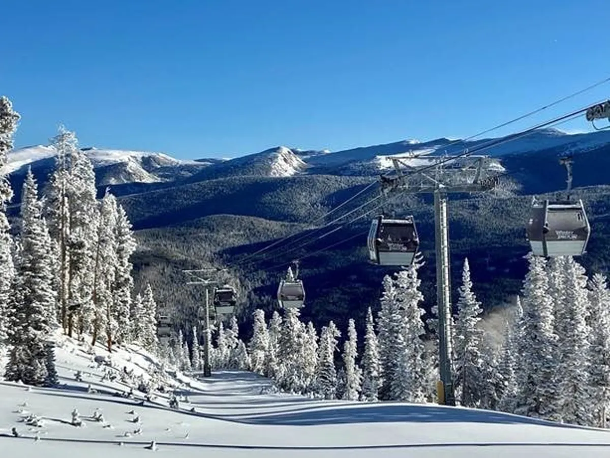 A scenic view of a ski lift gliding over snow-covered trees, with a majestic mountain range in the background and a clear blue sky overhead. The foreground features pristine winter slopes, inviting guests to enjoy skiing, snowboarding, and other winter sports.