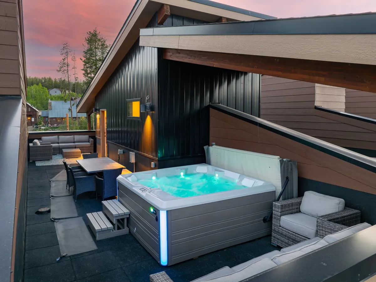 An inviting outdoor scene featuring a bubbling hot tub surrounded by comfortable seating and a large wooden dining table, set beneath sturdy wooden beams. The space is perfect for relaxation or social gatherings, with views of the surrounding natural beauty.