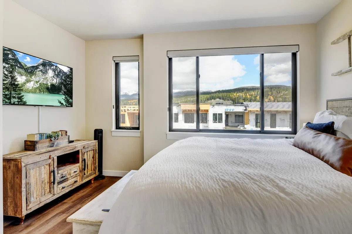 A serene bedroom with a comfortable bed dressed in textured bedding, a large window offering sweeping mountain views, and a wooden media console with a Smart TV. The calming décor incorporates subtle natural elements, creating a peaceful retreat.