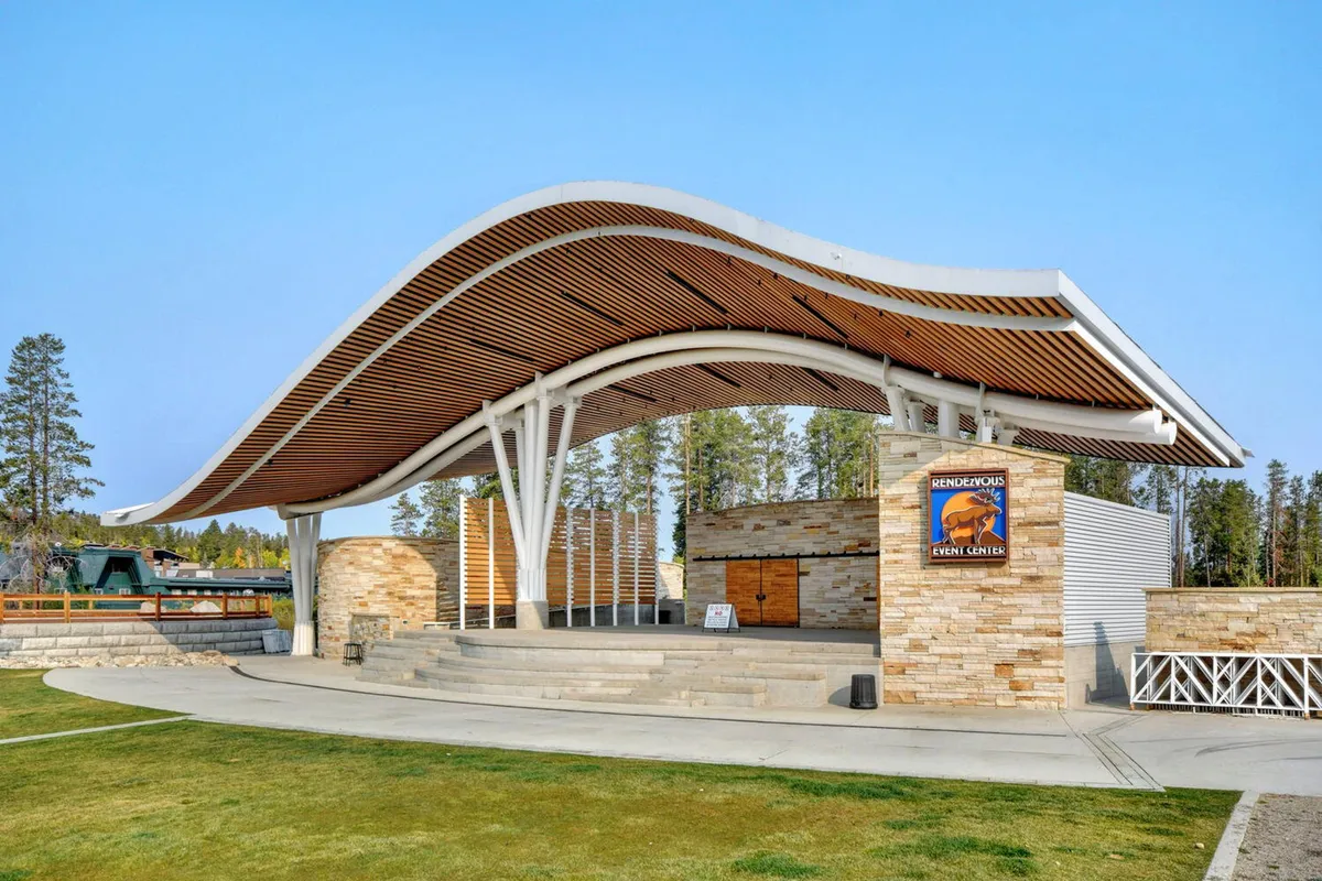 An exterior view of the Hideaway Park amphitheater featuring a unique architectural structure with a natural stone facade, surrounded by lush greenery. The open lawn invites gatherings and the space is designed for community events and concerts under the stars.
